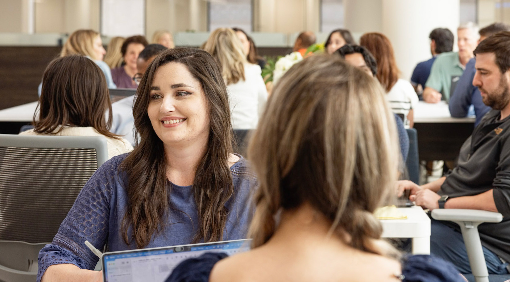 A woman with long dark hair and a blue top smiles while working on a laptop in an open office space filled with people. Others are engaged in various activities, including conversations and working at desks. The atmosphere appears collaborative and friendly.