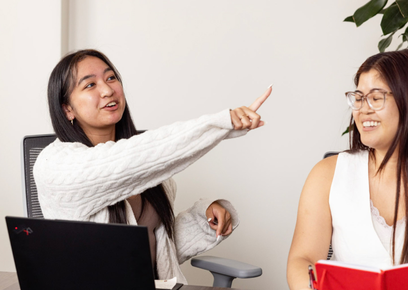 Two people are seated in an office setting, engaged in conversation. The person on the left, wearing a white sweater, is pointing upwards and appears to be speaking. The person on the right, wearing glasses and a sleeveless top, is smiling. A laptop is on the table.