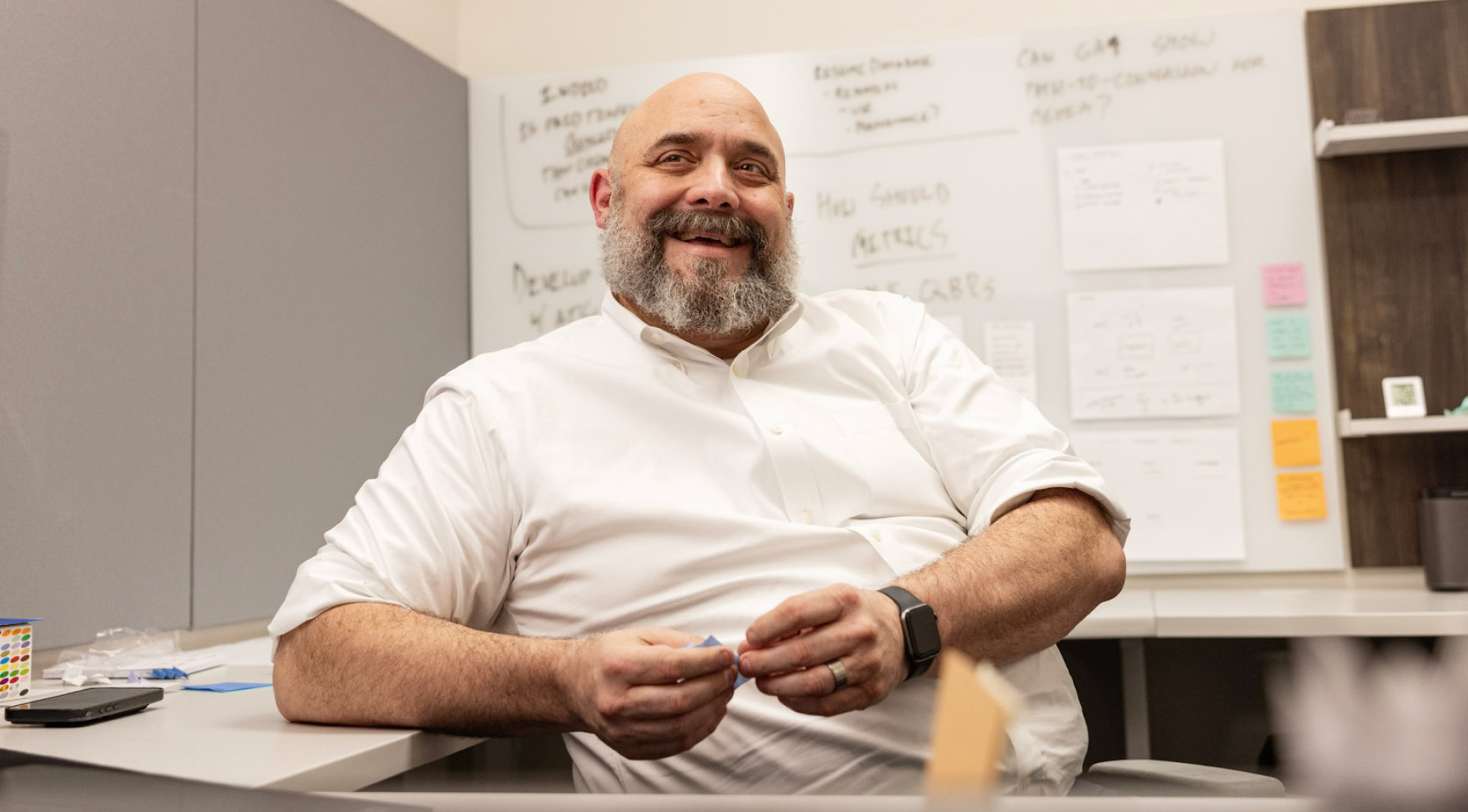 A man with a beard and wearing a white shirt is smiling while sitting at a desk in an office. Behind him, a whiteboard filled with notes and a shelf with assorted items are visible. The atmosphere appears casual and relaxed.