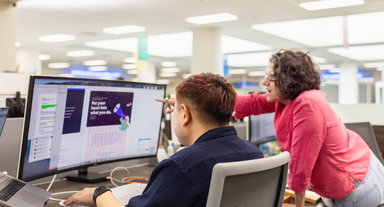 Two individuals are working at a desk with a large computer monitor displaying a design. A person in a red shirt is standing and pointing at the screen while talking to a seated person in a dark shirt. The office background is brightly lit with modern furnishings.