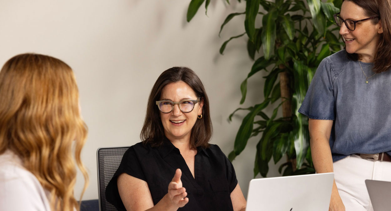 Three individuals engaged in a conversation in a modern office setting. One person in the center is speaking while gesturing with her hand, and two others are listening attentively. A laptop is open on the table, and a potted plant is visible in the background.